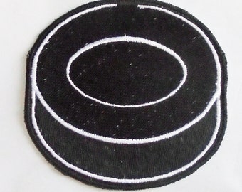May include: Black embroidered patch with a white outline. The patch is circular with a raised, oval-shaped centre. The design resembles a hockey puck. The patch is likely for clothing or accessories.