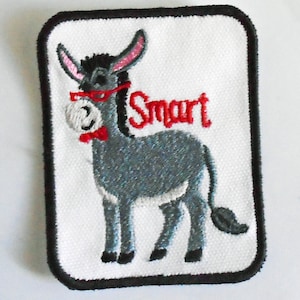 Könnte beinhalten: Ein weißer gestickter Aufnäher mit einem grauen Esel, der eine rote Brille und eine rote Schleife trägt. Der Esel steht auf einem weißen Hintergrund und das Wort "Smart" ist in Rot über dem Esel gestickt.