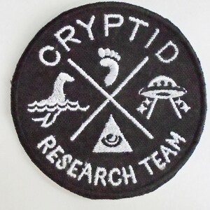 May include: Black and white embroidered patch with the text "Cryptid Research Team" in a circle around a cross with a footprint, a flying saucer, a sea monster, and a triangle with an eye inside.