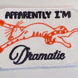 May include: White embroidered patch featuring an orange cat leaping in the air with the text "APPARENTLY I'M Dramatic" in black.