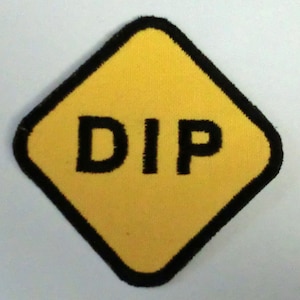 Puede incluir: Señal amarilla en forma de diamante con borde negro y la palabra "DIP" en letras negras.