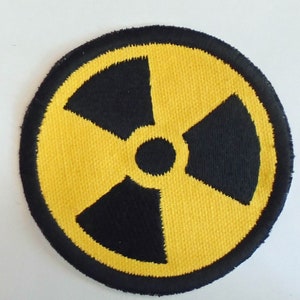 Op de afbeelding: Een gele en zwarte geborduurde patch met een stralingssymbool. Het symbool is een gestileerd klavertjevier met een zwarte cirkel in het midden.