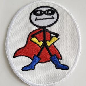 以下が含まれることがあります： 白い楕円形の刺繍パッチ。赤いマント、黄色の手袋、青いブーツをつけた棒人間スーパーヒーローが描かれている。