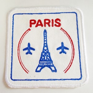 May include: White fabric patch with blue and red embroidery. The patch features a stylized Eiffel Tower, two airplanes, and the word "PARIS" in red.