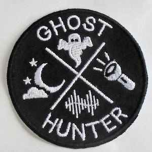 May include: Black and white embroidered patch with the text "Ghost Hunter" in a circle. The patch features a ghost, a crescent moon, a flashlight, and sound waves.