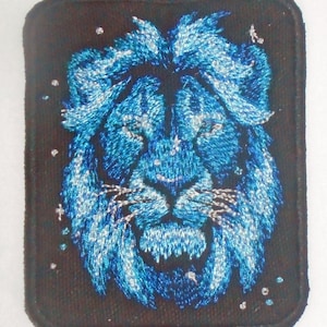 Puede incluir: Parche bordado azul con la cabeza de un león sobre un fondo negro. La melena del león está detallada con costuras blancas.