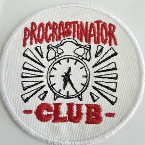 May include: White, circular embroidered patch with the words "PROCRATINATOR CLUB" in red. A black and white alarm clock design is in the center, with radiating lines. The patch has a white border.