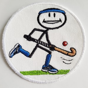 Op de afbeelding: Een witte geborduurde patch met een stokfiguur die veldhockey speelt. De stokfiguur draagt een blauwe hoofdband, blauwe sokken en blauwe schoenen. De stokfiguur houdt een bruin en zwart hockeystick vast en een rode bal is in de lucht.