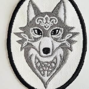 May include: A white embroidered patch with a black border, featuring a grey wolf's face with intricate details and a Celtic-style design.