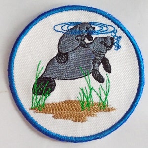 May include: Embroidered patch featuring a gray manatee with a baby manatee on its back, swimming in blue water with green grass and brown sand below. The patch has a blue border.