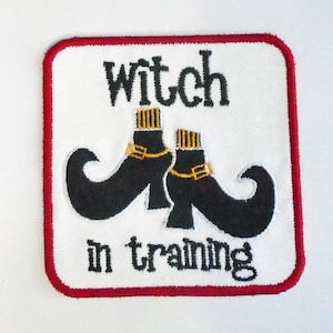 May include: Square white fabric with a red border, embroidered with the words "witch in training" in black. Below the text are two black witch boots with gold buckles and accents.