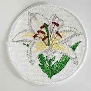 Könnte beinhalten: Weißer gestickter Lilienblumen-Patch mit grünen Blättern und roten Staubfäden. Der Patch ist rund und hat einen weißen Rand.