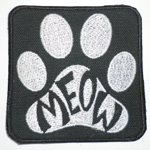 May include: Square black patch with a white paw print design. The word "MEOW" is embroidered in white within the paw print. The patch has a black border and is likely an iron-on or sew-on patch.