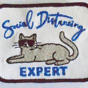 Könnte beinhalten: Weißer gestickter Aufnäher mit einer Comic-Katze, die eine Sonnenbrille trägt, und dem Text "Social Distancing Expert" in Blau.