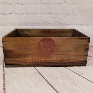 May include: A vintage wooden crate with a rectangular shape. The crate is made of aged wood with visible grain and knots. Red text on the front reads "Swift's Premium" in a circular design. The crate sits on a light-colored wooden surface.