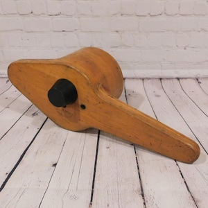 May include: A vintage wooden shoe last, featuring a light brown finish with a black knob. The shoe last has a rounded heel and a long, flat handle, ideal for shaping footwear. The object is positioned on a light-colored wooden surface.