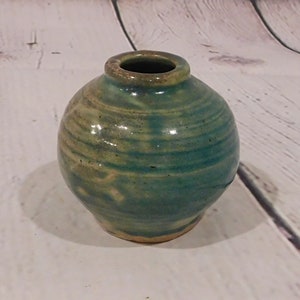 May include: A small, round ceramic vase with a narrow opening. The vase is primarily teal and green, with a swirling pattern and a light brown rim. The handmade pottery has a rustic, organic aesthetic.