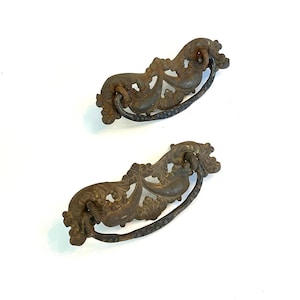 May include: Two matching drawer pulls featuring an ornate, weathered bronze-toned metal backplate with intricate scrollwork and a curved, textured drop handle. The metal surfaces show signs of oxidation and age, with a dark, mottled patina.