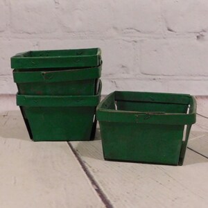 May include: A collection of small, square, green wooden berry baskets. The baskets are stacked and arranged on a light-colored wooden surface. The baskets have a rustic, vintage appearance.