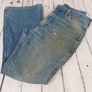 May include: Light blue denim jeans with a faded appearance and visible wear. The jeans have a classic five-pocket design, a button and zipper closure, and a straight leg. The fabric shows signs of use, with some discoloration and stains.