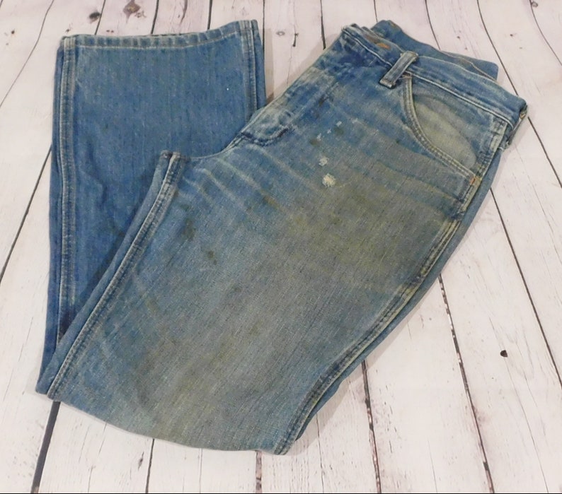 May include: Light blue denim jeans with a faded appearance and visible wear. The jeans have a classic five-pocket design, a button and zipper closure, and a straight leg. The fabric shows signs of use, with some discoloration and stains.