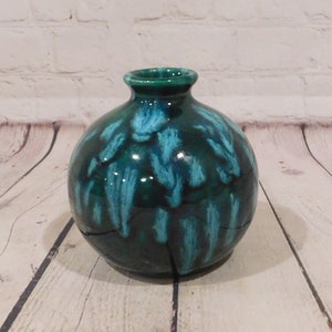 May include: A round ceramic vase with a dark green base and turquoise drip glaze. The vase has a small opening at the top and a crack running down the side. The vase is sitting on a wooden surface.