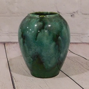 May include: A rounded ceramic vase featuring a glossy glaze with a mottled blend of deep forest green, emerald, and dark teal tones. The surface exhibits subtle variations in color intensity and texture, creating a fluid, organic appearance.