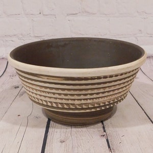 May include: A decorative ceramic bowl with a dark brown interior and a cream-colored rim. The exterior features horizontal bands with a carved, scalloped design. The bowl sits on a pedestal base, showcasing a rustic, handcrafted aesthetic.