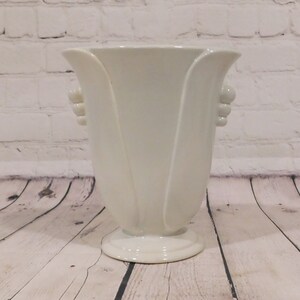 May include: A glossy off-white ceramic vase featuring a flared rim and a pedestal base. The body of the vase has vertical, petal-like raised panels and decorative three-tiered bead accents on each side, creating a structured, symmetrical silhouette.