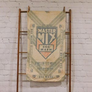 May include: Vintage feed sack for "Master Mix" egg mash, with text and graphics in orange, blue, and green. The sack is suspended from a wooden ladder. The text includes "100 LBS NET" and "Millen Feed Mills".
