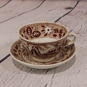 May include: A vintage teacup and saucer set. The cup and saucer are cream-colored with a brown transferware design. The cup features floral and landscape patterns. The saucer has a matching design. The set is likely made of ceramic.