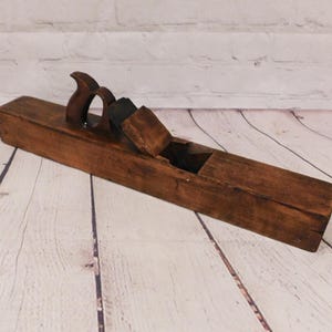 May include: A vintage wooden hand plane, showcasing a rich, aged brown color. The plane features a curved handle and a blade housing, indicative of its woodworking function. The tool rests on a light-colored wooden surface, suggesting a workshop setting.