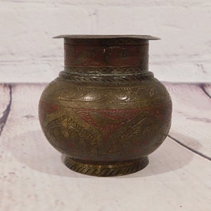 May include: An antique brass pot with a bulbous body and a flared rim. The pot features intricate etched floral patterns and red accents. The base has a decorative, ridged design. The pot is likely used for storage or display.
