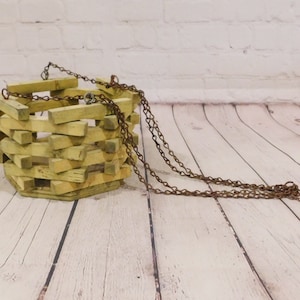 May include: A hanging planter constructed from light yellow wooden slats arranged in a stacked, log-cabin style pattern. The structure is hexagonal and features a rusted metal chain attached to the top for suspension, creating a rustic appearance.