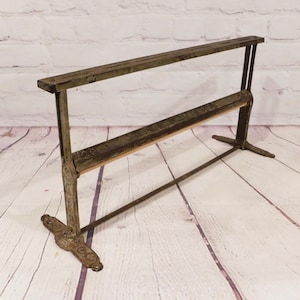 May include: A vintage metal paper dispenser with a weathered green finish and ornate cast iron feet. The structure features two vertical supports, a horizontal bar for paper rolls, and a serrated cutting edge for tearing paper sheets cleanly.