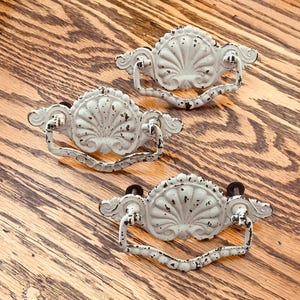 May include: Three off-white metal drawer pulls featuring a scalloped shell design with a distressed finish. Each handle has a curved, hinged drop pull attached to a decorative backplate with ornate scrollwork details and dark metal hardware.