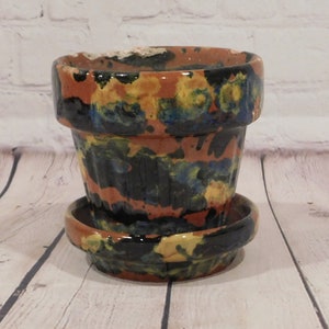 May include: A ceramic flower pot with a matching saucer. The pot is painted with a mix of colors, including black, orange, yellow, and blue. The pot has a classic shape with a slightly flared rim. The saucer is round and has a matching color scheme.