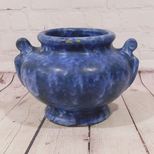 May include: A blue ceramic vase with a speckled pattern. The vase has two handles and a pedestal base. The interior is a darker shade of blue. The vase is approximately 8 inches in diameter and 6 inches tall.