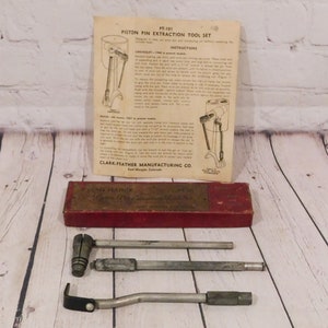 May include: A vintage piston pin extraction tool set, including a red box, instruction sheet, and several metal tools. The instruction sheet is labeled "PT-101 Piston Pin Extraction Tool Set" and includes diagrams. The box is labeled "Clark-Feather".