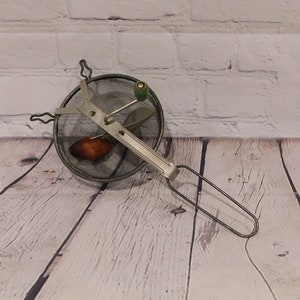 May include: A vintage food mill with a metal mesh bowl, a silver-toned handle, and a wooden knob. The mill has a silver-toned blade and a brown wooden piece inside the bowl. The handle is a loop shape.