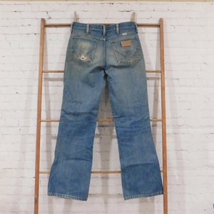 May include: Light blue denim jeans with a worn appearance, hanging on a wooden ladder. The jeans have a classic five-pocket design, a brown leather patch, and a distressed look with a hole in the back pocket.