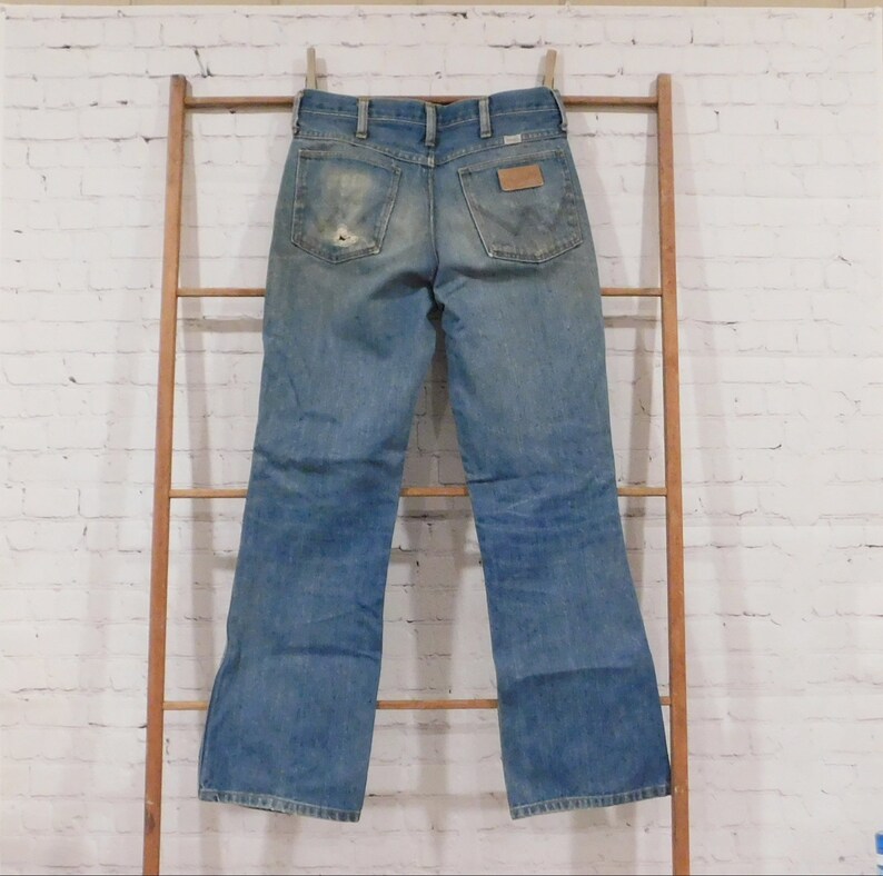May include: Light blue denim jeans with a worn appearance, hanging on a wooden ladder. The jeans have a classic five-pocket design, a brown leather patch, and a distressed look with a hole in the back pocket.