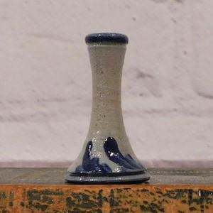 May include: A small, handmade ceramic vase with a speckled, light gray body and a dark blue rim and base. The vase features dark blue wave-like designs near the base. The vase is approximately 5 inches tall.