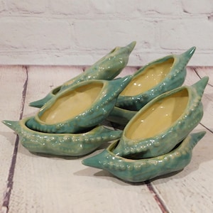 May include: A collection of five ceramic pea pod dishes featuring a glossy teal exterior with raised, textured bumps and a smooth, pale yellow interior. Each dish is shaped like an elongated, pointed pod, creating a cohesive set of vessels.