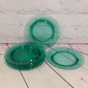 May include: A set of four translucent emerald green glass plates featuring a smooth center and a textured, patterned rim. The plates are arranged with one standing upright in the back, a stack of two on the left, and one lying flat on the right.