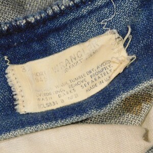 May include: Close-up of blue denim jeans with a white tag. The tag reads "WRANGLER" and includes washing instructions and size information. The jeans are slightly faded, showing signs of wear.
