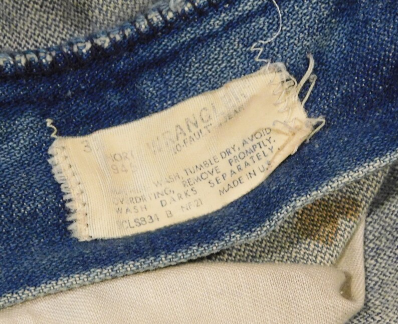 May include: Close-up of blue denim jeans with a white tag. The tag reads "WRANGLER" and includes washing instructions and size information. The jeans are slightly faded, showing signs of wear.