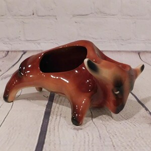 May include: A glossy ceramic planter shaped like a bull in a charging stance. The bull is colored in shades of brown and tan with black accents on the hooves, ears, and face. It features a hollow opening on its back for holding small items.