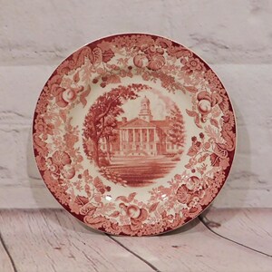 May include: A round, antique-style plate with a red and white floral and fruit pattern. The center features a red illustration of a building with a tower, surrounded by trees. The plate has a scalloped edge.