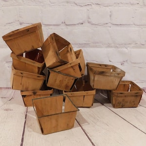 May include: A collection of square wooden berry baskets with a light brown finish and dark metal rims. The baskets are stacked in a pile, showing their open tops and thin wooden slats. Each basket has a simple, functional design for holding items.